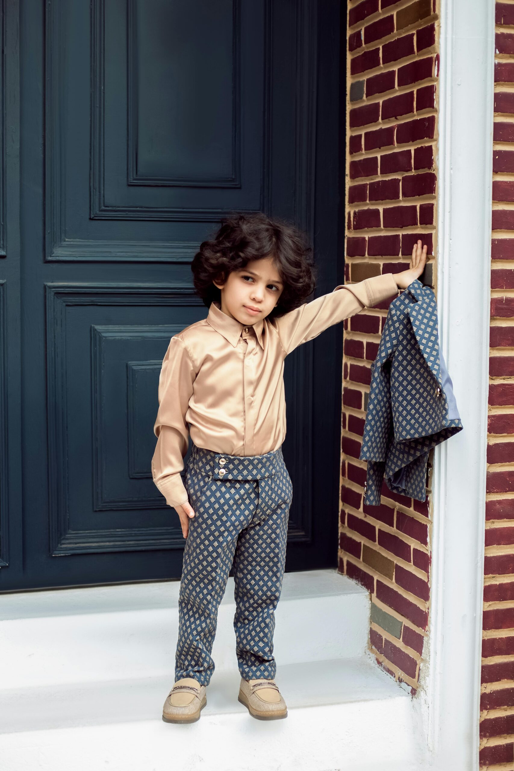 A young child dressed impeccably in a fashionable outfit poses confidently by a doorway in an urban setting.